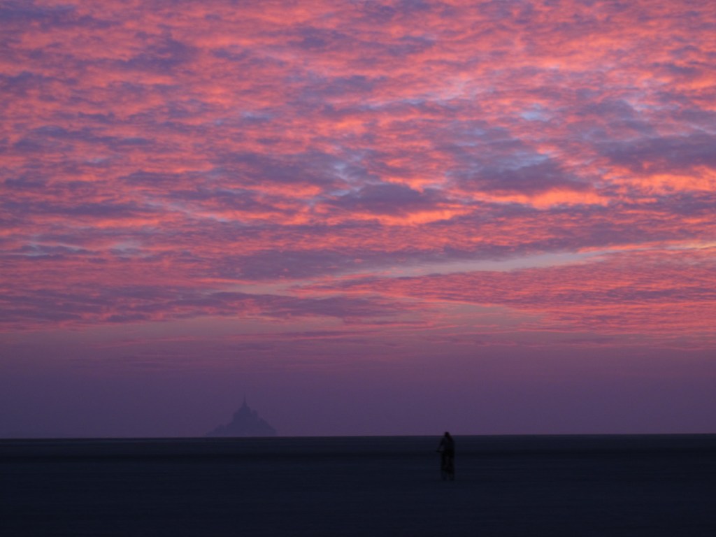 Sonnenuntergang hinter Le Mont-Saint-Michel, Frankreich, mit einsamen Radfahrer auf dem Watt.