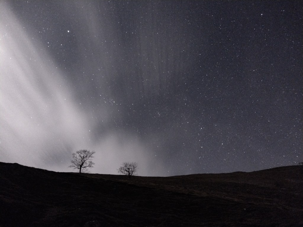 Die ersten Wolken eines großes Wolkengebiets, von einer dünnen Mondsichel angeleuchtet, ergänzen die Komposition vom Sternenhimmel über einem Steinbruch mit kahlen Bäumen