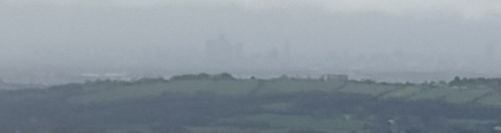 Blick von der Snake Road nach Manchester mit viel digitalem Zoom. Man kann gerade noch die Umrisse der Wolkenkratzer erkennen.